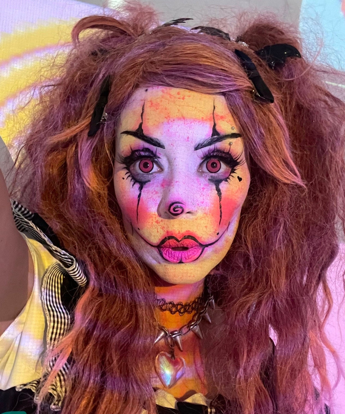 A close-up of a person with elaborate horror clown makeup, featuring bright pink lips, black painted eyes, and a spiral nose, wearing a messy orange-brown wig with black hair clips and a spiked choker.